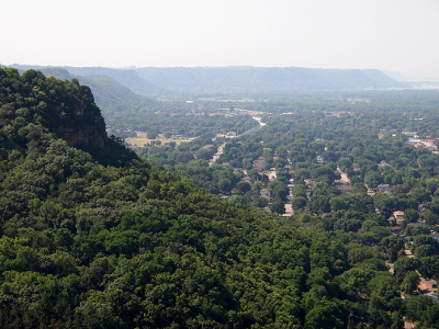 [The bluffs, completely covered in greenery, rise sharply from the river-level plain which contains roads and houses amids the mature trees. The bluffs give the effect of an amphitheater as they encircle the lower level on three sides.]